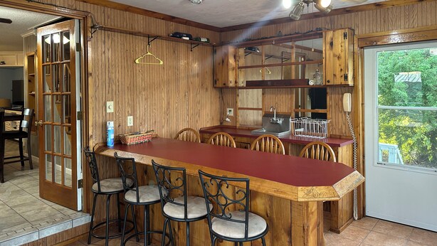 A kitchen with wooden cabinets, a bar area with stools, a ceiling fan, and a window with a view of greenery. - Fishing camp in Falgout Canal Marina! (theriot)
