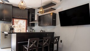 A modern kitchen with dark cabinetry, a central island with bar stools, pendant lighting, and a flat-screen TV mounted on the wall. - Cozy Studio Apartment.  Pet Friendly! (Sayulita)
