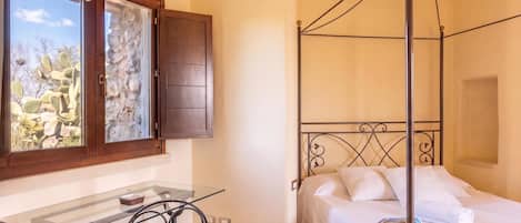 A bedroom with a four-poster bed, a glass table with a chair, a window with a view of cacti, and a door with a wooden frame.