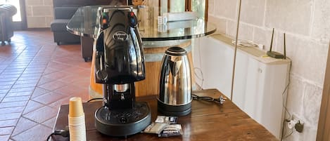 Una cafetera, un hervidor y una pila de tazas sobre una mesa de madera en una habitación con suelo de baldosas y una ventana.