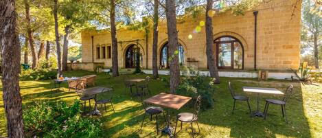 Un patio al aire libre con mesas y sillas, rodeado de árboles y un edificio de piedra con ventanas arqueadas.
