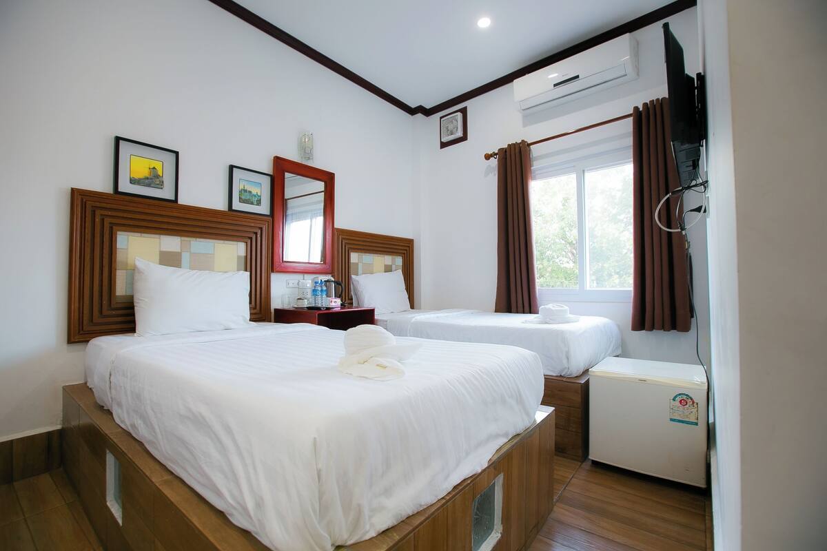 Une chambre d’hôtel avec deux lits, une petite table avec une lampe, un réfrigérateur et une fenêtre avec des rideaux.