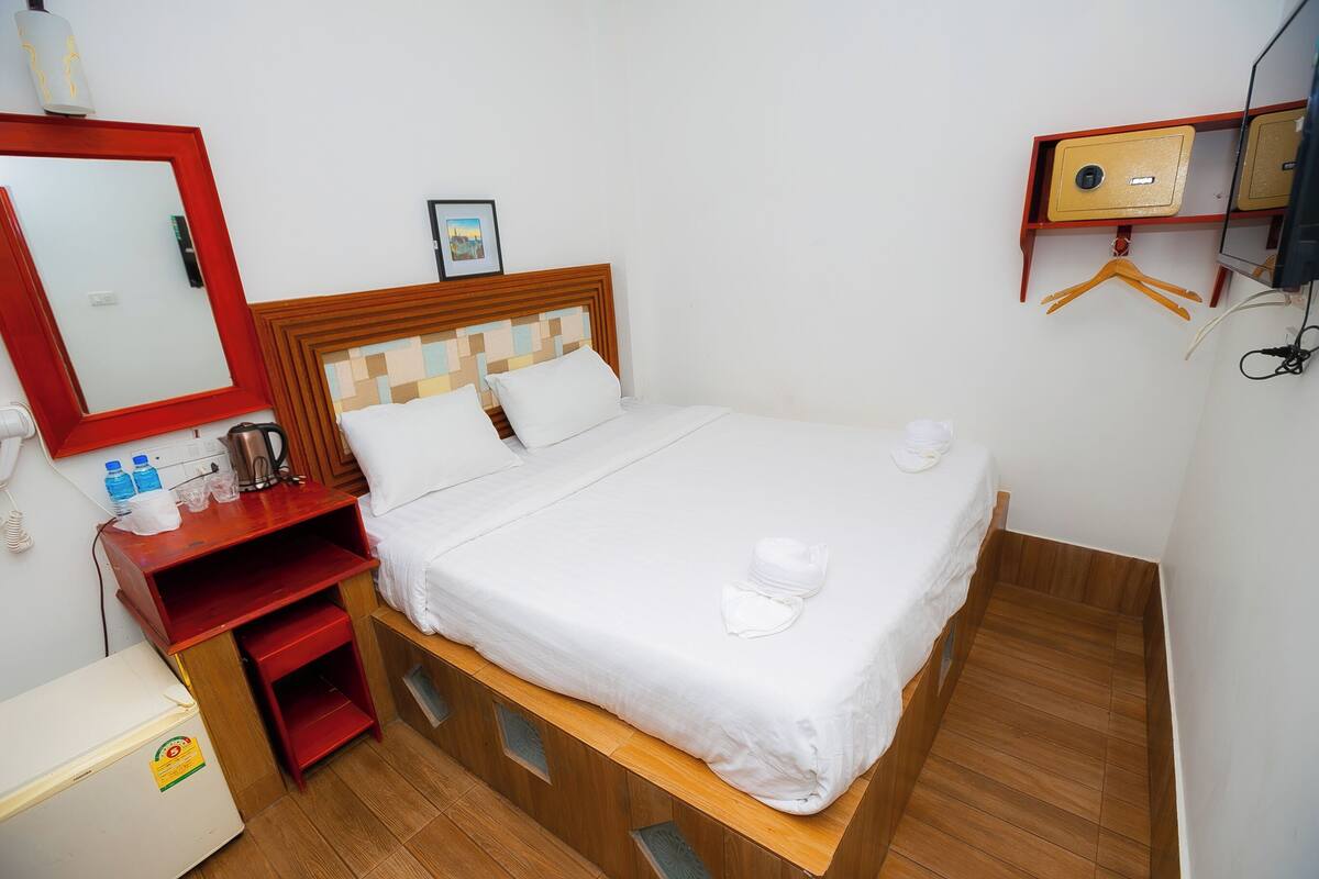 Un lit bien fait, avec du linge de lit blanc, une tête de lit en bois, une petite table de chevet avec des articles de toilette et une table de nuit rouge.