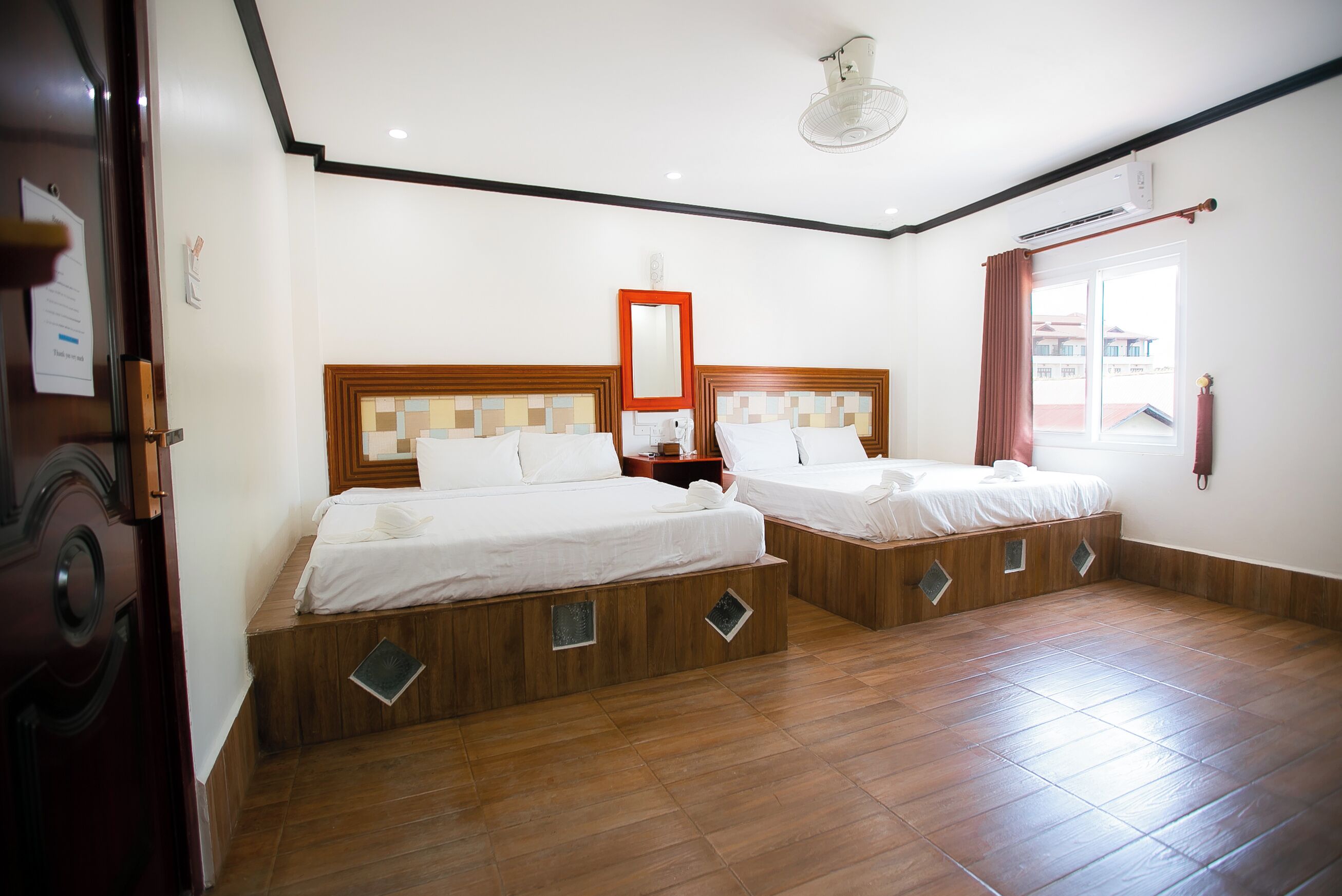 A hotel room with two beds, wooden headboards, a mirror, a window with curtains, and a ceiling fan.