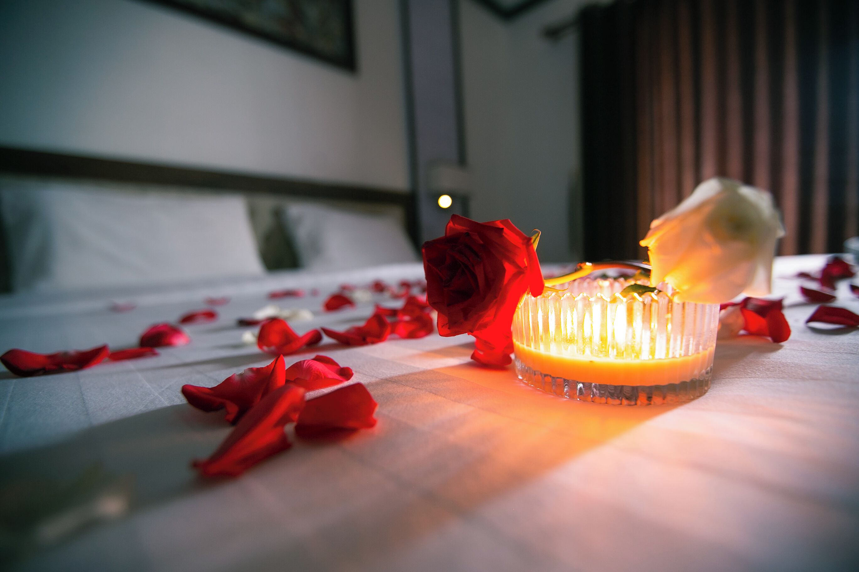 A bed with a white bedspread, a red rose, and a lit candle on a table.