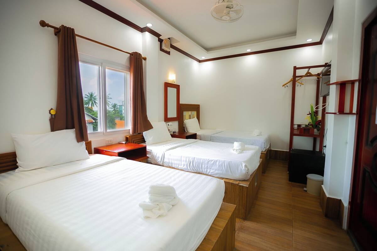 Une chambre d’hôtel avec deux lits, un plancher en bois, une fenêtre avec des rideaux, une table de chevet, un miroir et une plante suspendue.
