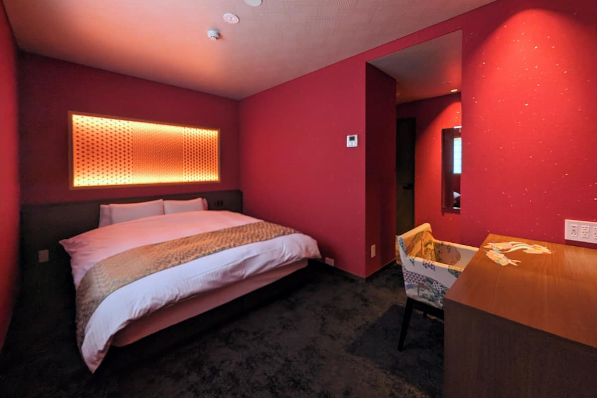 Une chambre d’hôtel avec un grand lit, une tête de lit ornée d’un panneau lumineux texturé, un bureau en bois et une chaise à motifs.