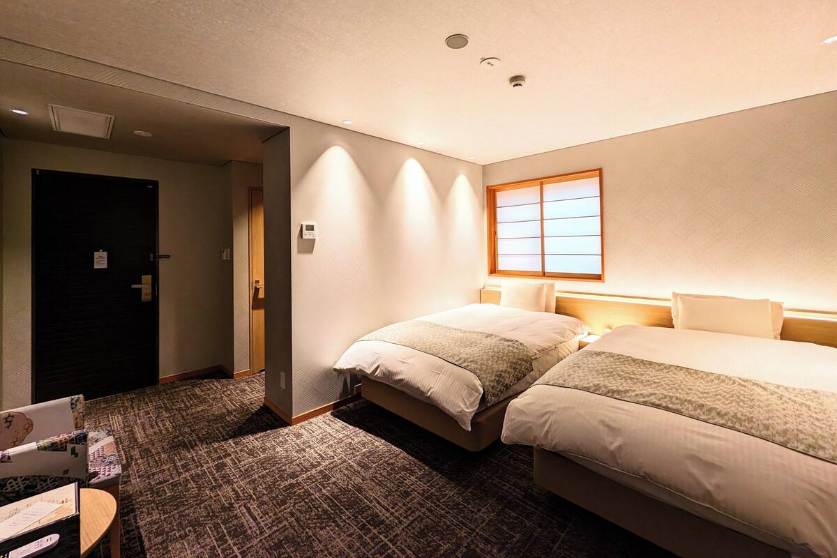 Une chambre d’hôtel avec deux lits, un sol recouvert de moquette, une porte en bois et une fenêtre avec des stores.