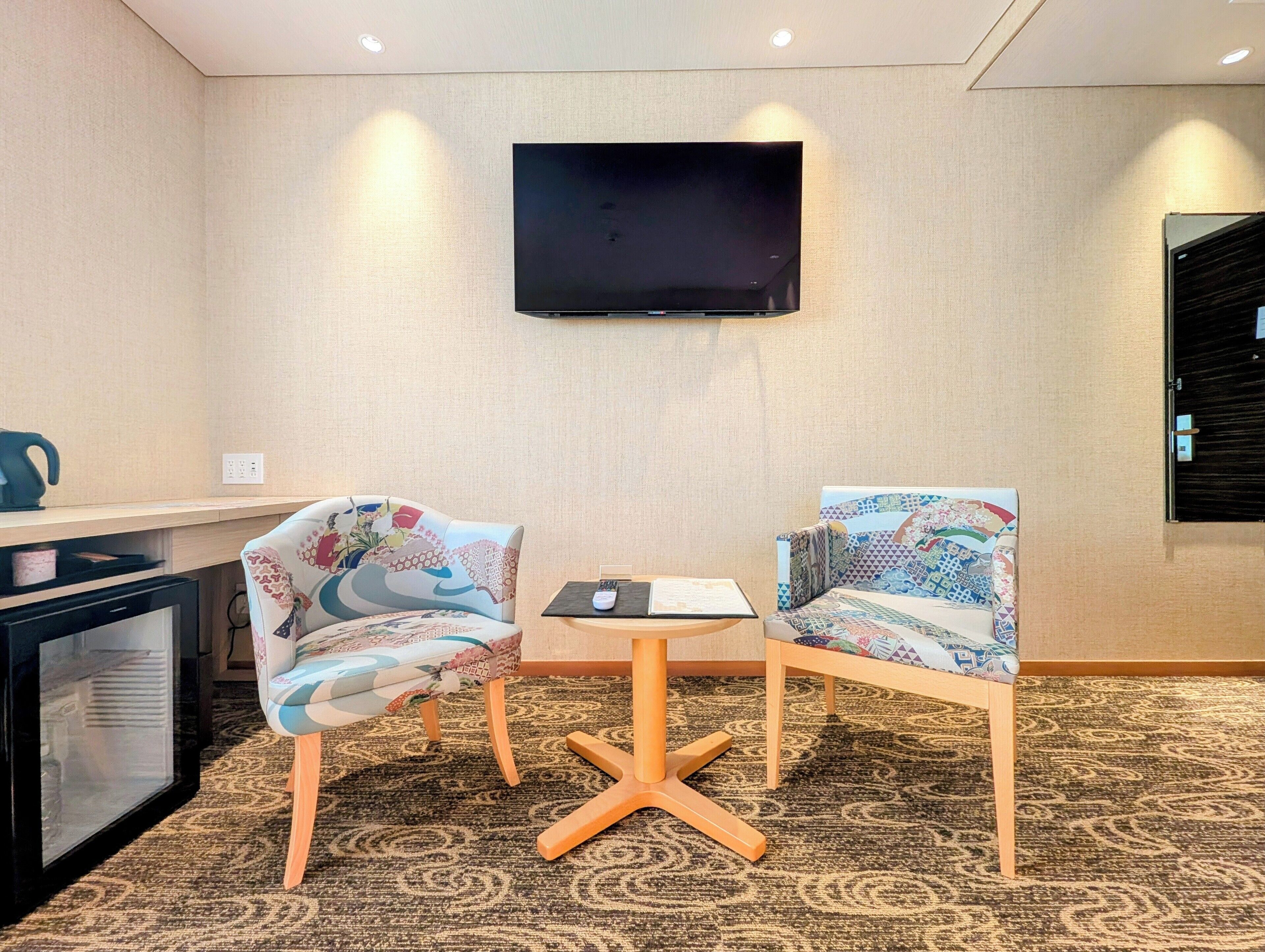 A hotel room with a flat-screen TV mounted on the wall, two patterned chairs around a small table, and a minibar to the left.