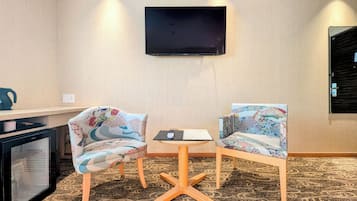 A hotel room with a flat-screen TV mounted on the wall, two patterned chairs around a small table, and a minibar to the left.