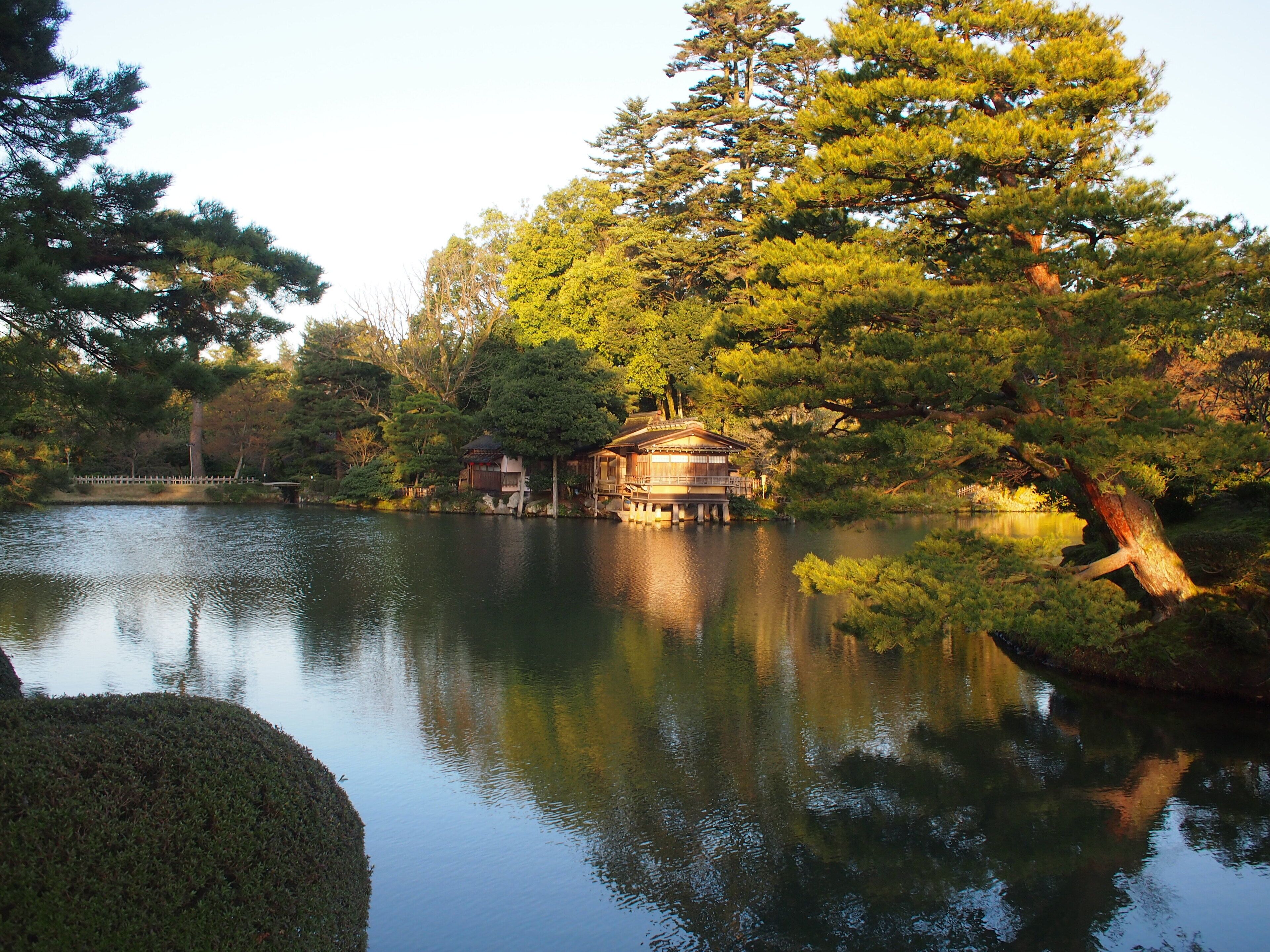 A traditional Japanese-style building with a wooden structure and a thatched roof, situated by a calm lake surrounded by lush greenery and trees.