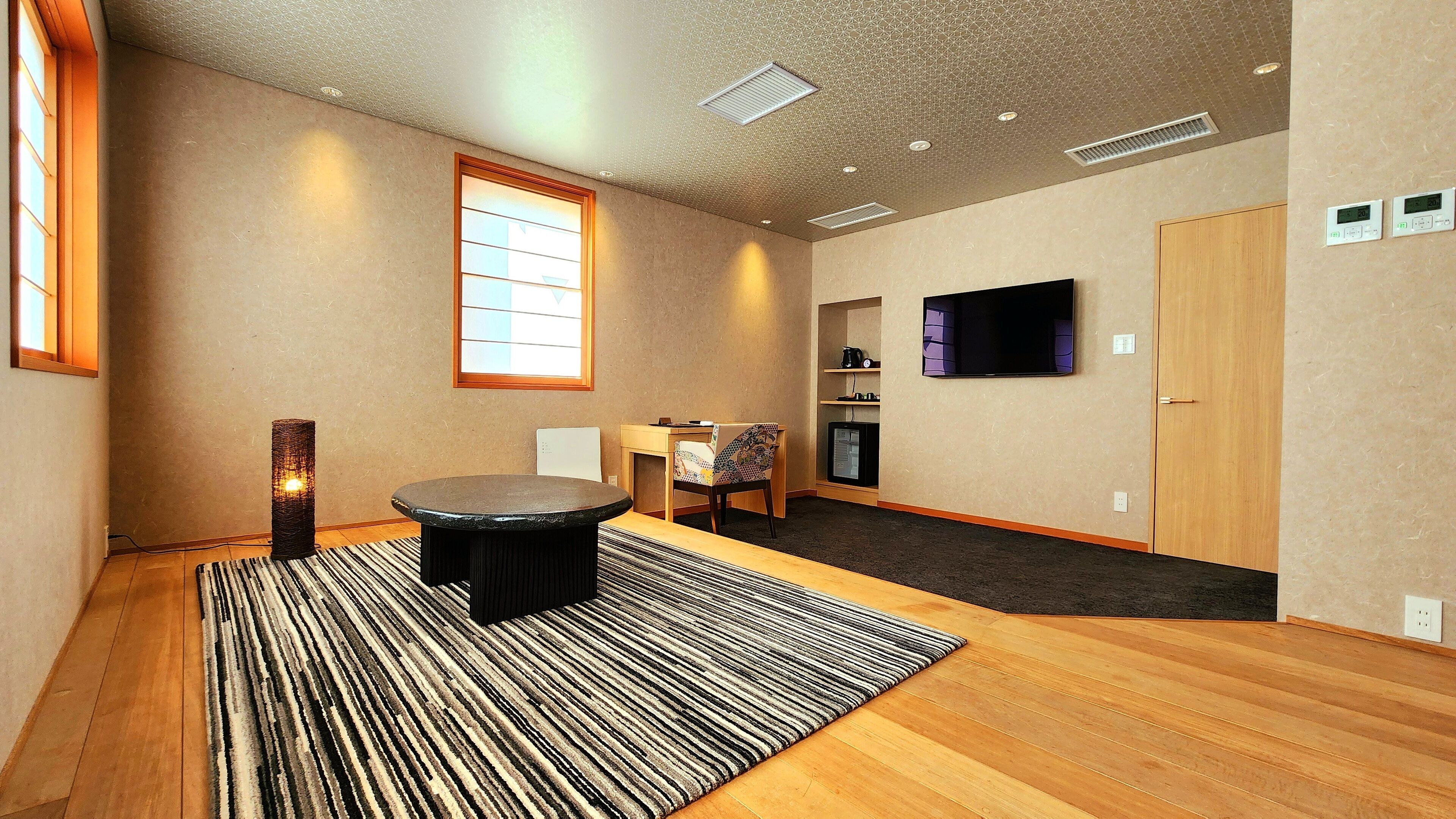 A room with a wooden floor, a round table, a patterned rug, a television, and a window with blinds.