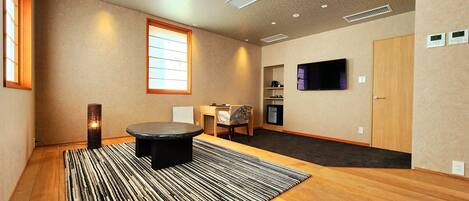 A room with a wooden floor, a round table, a patterned rug, a television, and a window with blinds.