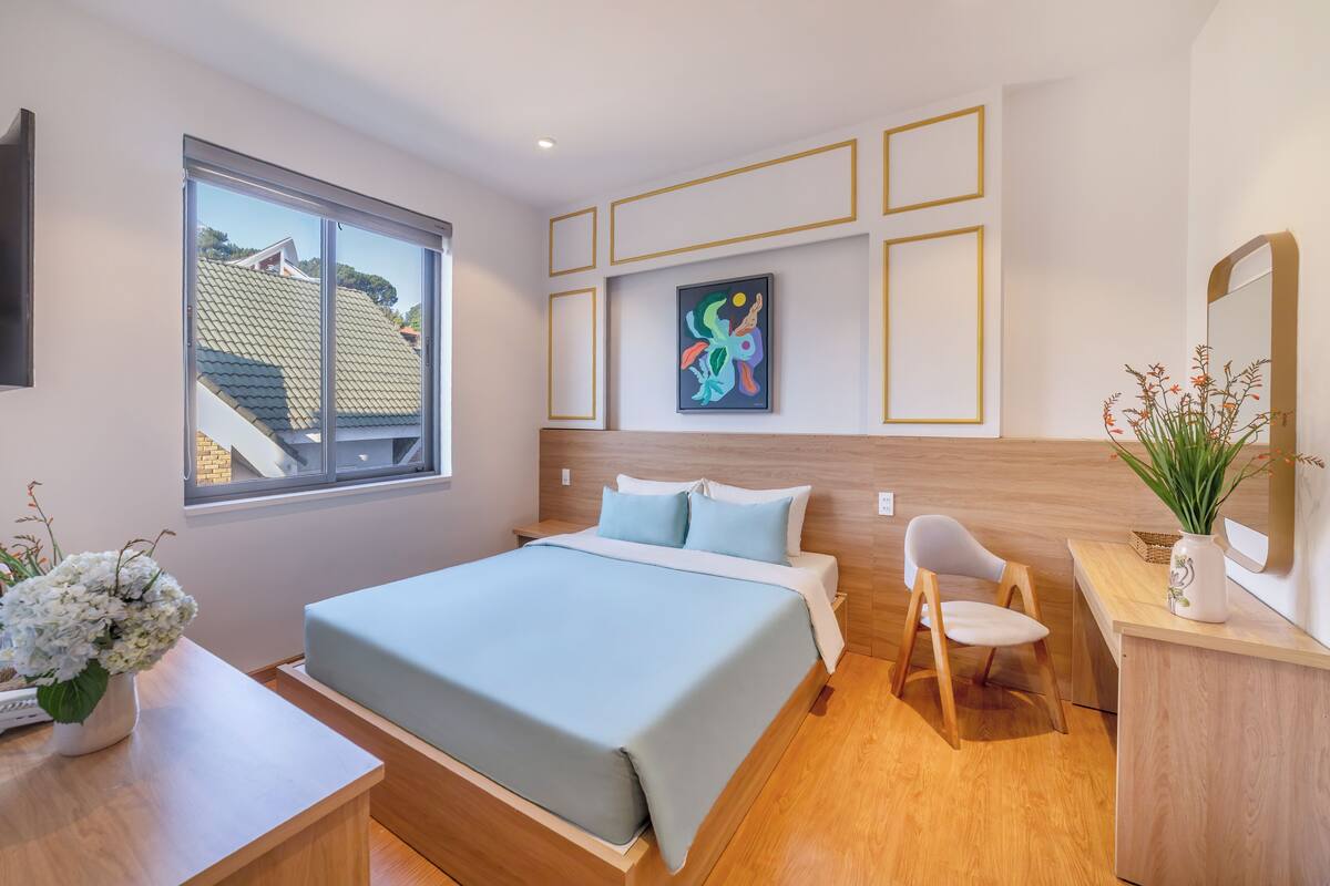 A modern bedroom with a large bed, a desk, a chair, a window, and a painting on the wall.
