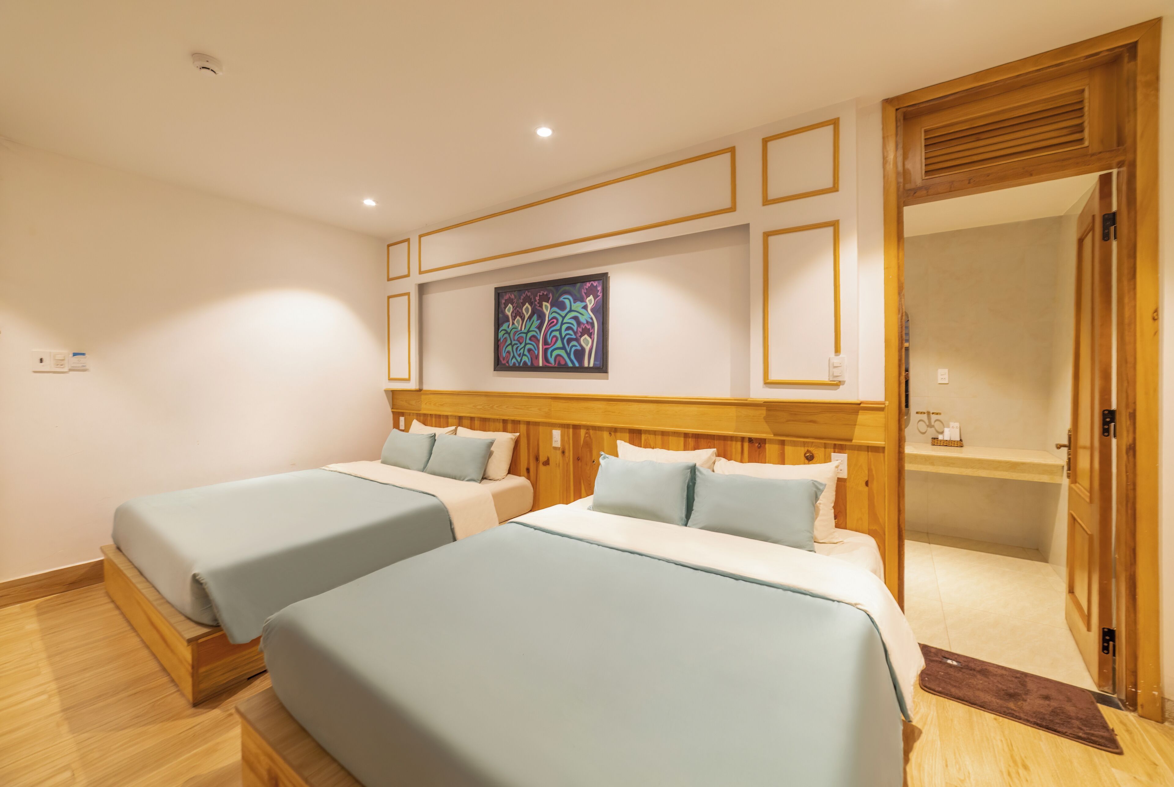 A hotel room with two beds, wooden headboards, a colorful artwork on the wall, and a wooden door leading to a bathroom.