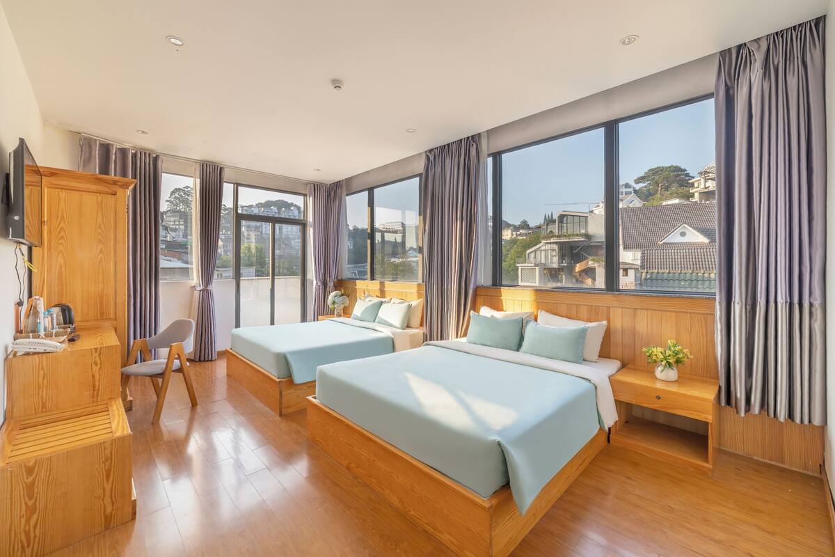 A hotel room with two beds, a wooden desk, a chair, and large windows offering a city view.