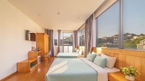 A hotel room with two beds, a wooden desk, a TV, and large windows offering a city view. - Pham Gia Villa &Hotel (Xuan Huong - Da Lat)