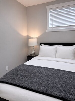A bedroom with a bed, a nightstand, a lamp, and a window with blinds. - Modern NW Retreat - Banff access (Calgary)