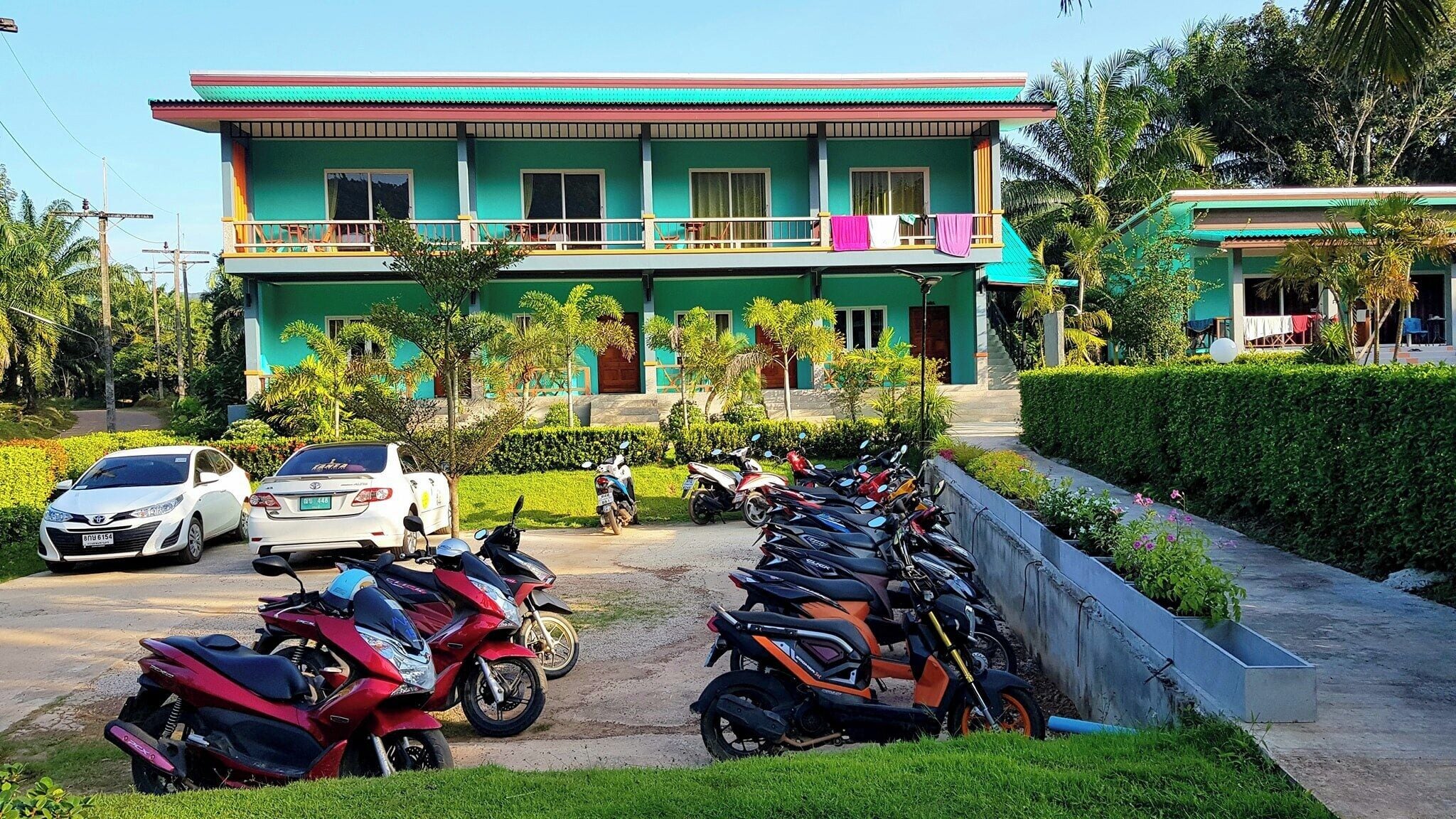 Een gebouw met twee verdiepingen, een groene gevel, een balkon met een paarse handdoek en een rij geparkeerde scooters en auto's.