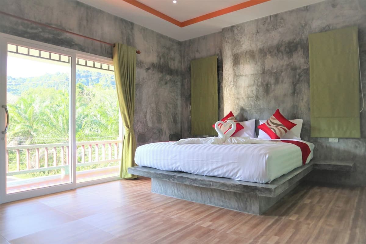 A bedroom with a bed on a platform, large sliding glass doors to a balcony, and a view of greenery outside.