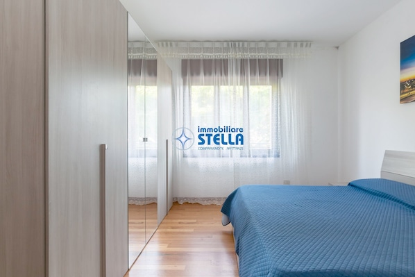 Une chambre à coucher avec un lit, une armoire, une fenêtre avec des rideaux légers et un tableau au mur.