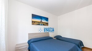 A bedroom with two beds, a nightstand, a drawer unit, and a wall-mounted picture of the Eiffel Tower. - Nausicaa Apartment  *few meters from the sea* (Jesolo)