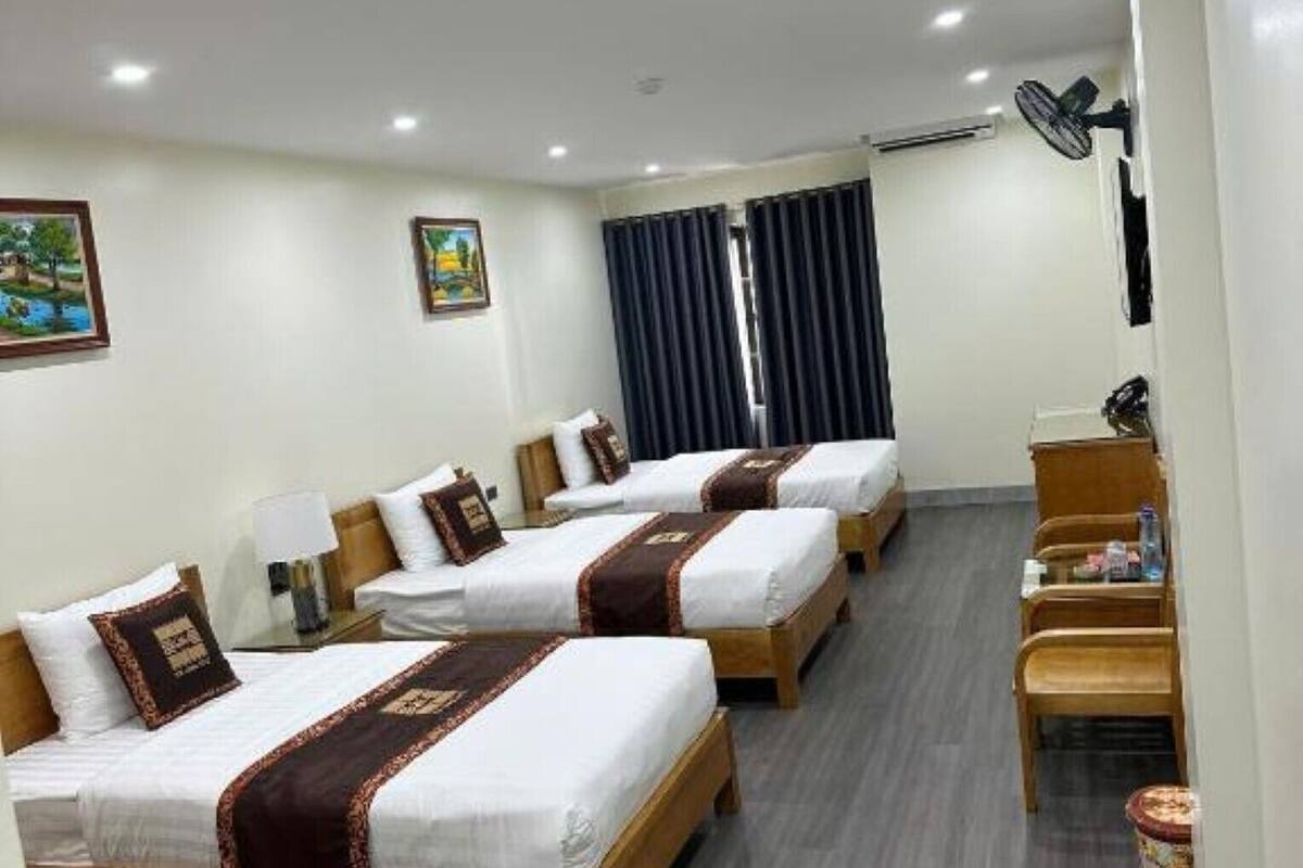 Habitación de hotel con tres camas, un escritorio, una silla, un cubo de basura y un ventilador de techo.