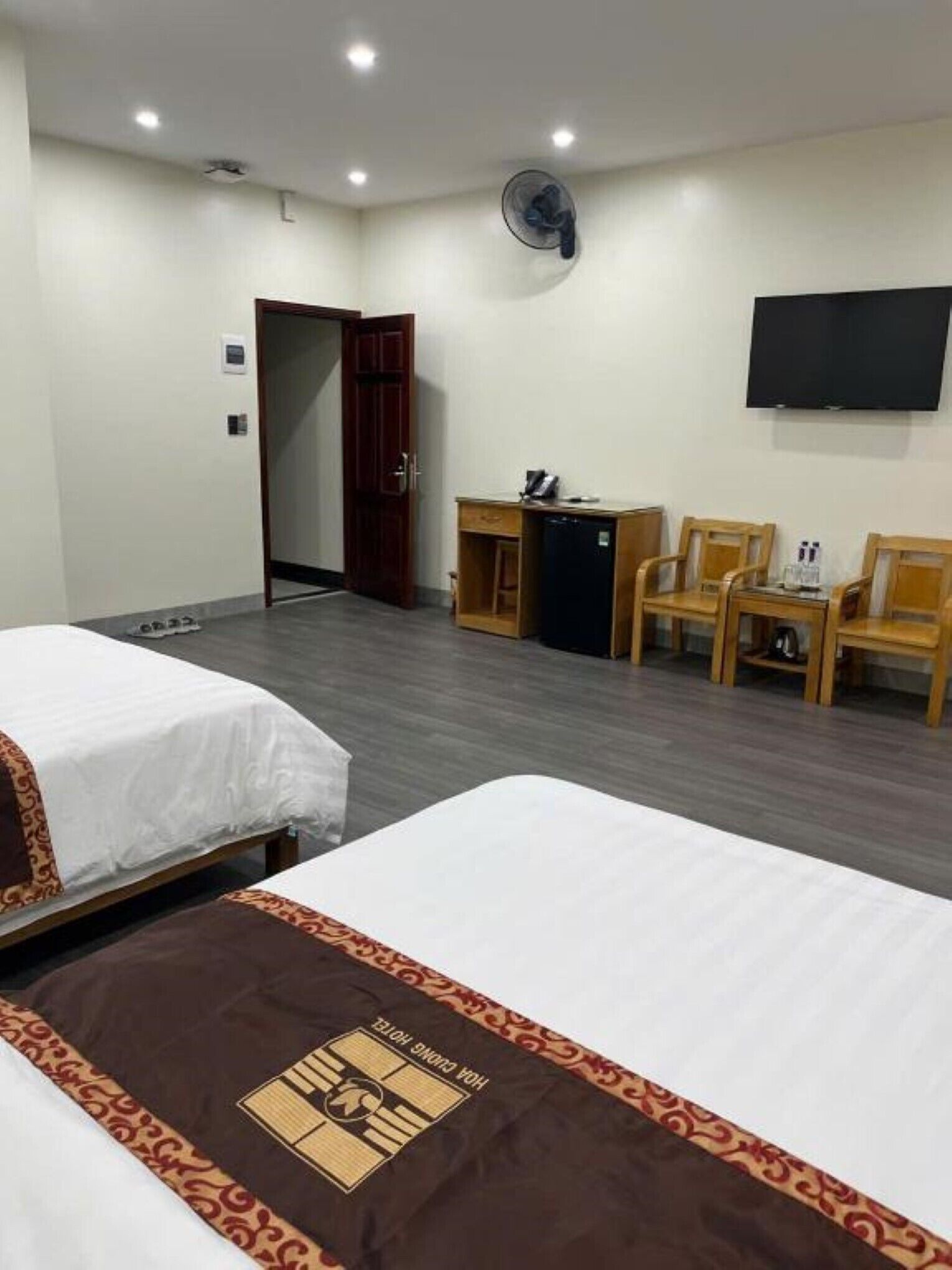 A hotel room with two beds, a desk, a chair, a TV, and a fan.
