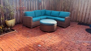Un patio con un sofá y reposapiés de mimbre, rodeado de una valla de madera y plantas en macetas.