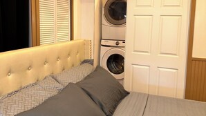 Un dormitorio con cabecera capitonada, cama con ropa de cama gris, lavadora y una puerta blanca.