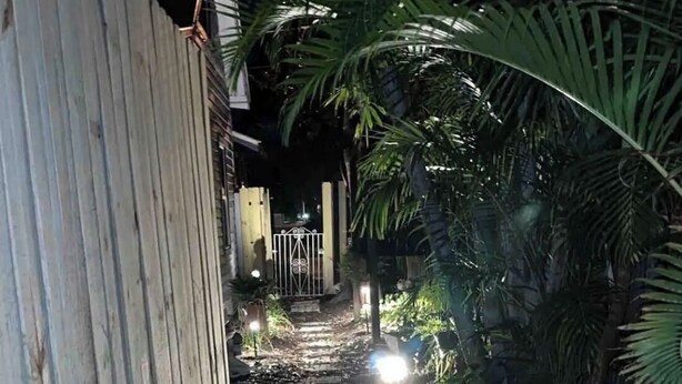 Un sendero estrecho que conduce a una entrada con puerta, rodeado de abundante follaje y una cerca de madera.