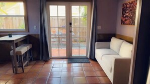 Una habitación con sofá blanco, una pequeña mesa con banqueta, una ventana y una puerta que da a un patio.
