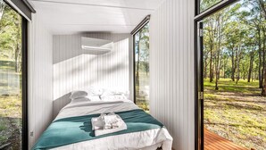 Un dormitorio moderno con cama, ropa de cama blanca, una manta turquesa y vistas a un bosque a través de amplios ventanales.