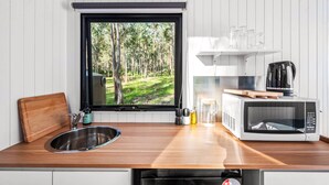 Una cocina con vista al bosque por una ventana, un lavamanos de acero inoxidable, un microondas blanco y una tabla de cortar de madera.