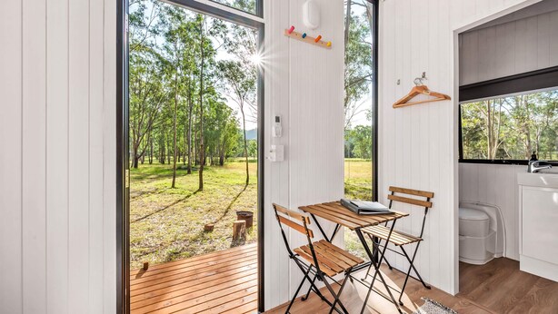 Una habitación con una mesa y sillas de madera, una ventana con vistas a árboles, un perchero y una pequeña cocina.