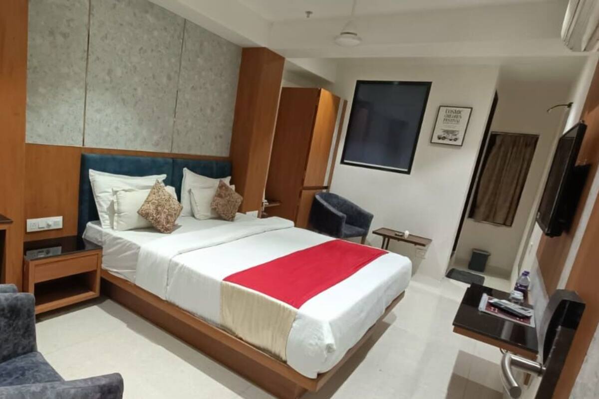 A hotel room with a large bed, wooden headboard, a desk with a chair, a small table, and a TV.