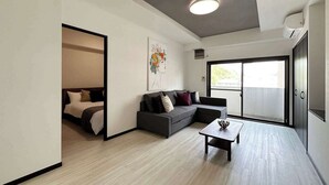 A modern living room with a grey sofa, a small coffee table, and a bed in the adjacent room. - 2Br Apt for 10ppl Very Good Location (Minami Ward, Hiroshima)