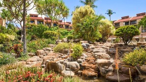 A landscaped garden with a rock pond, palm trees, and a building with red roofs in the background. - Walk to Beach | Pools, Pickleball, Kihei (Kihei)