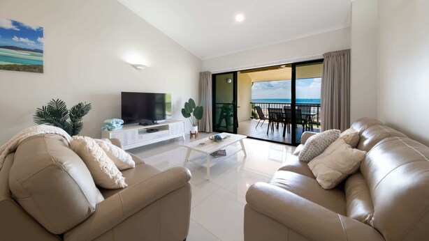 A modern living room with a sofa set, a coffee table, and a TV. There is a balcony with a view of the sea.