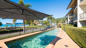 A swimming pool with a clear blue water, surrounded by a paved walkway, a black metal fence, and a large umbrella providing shade.