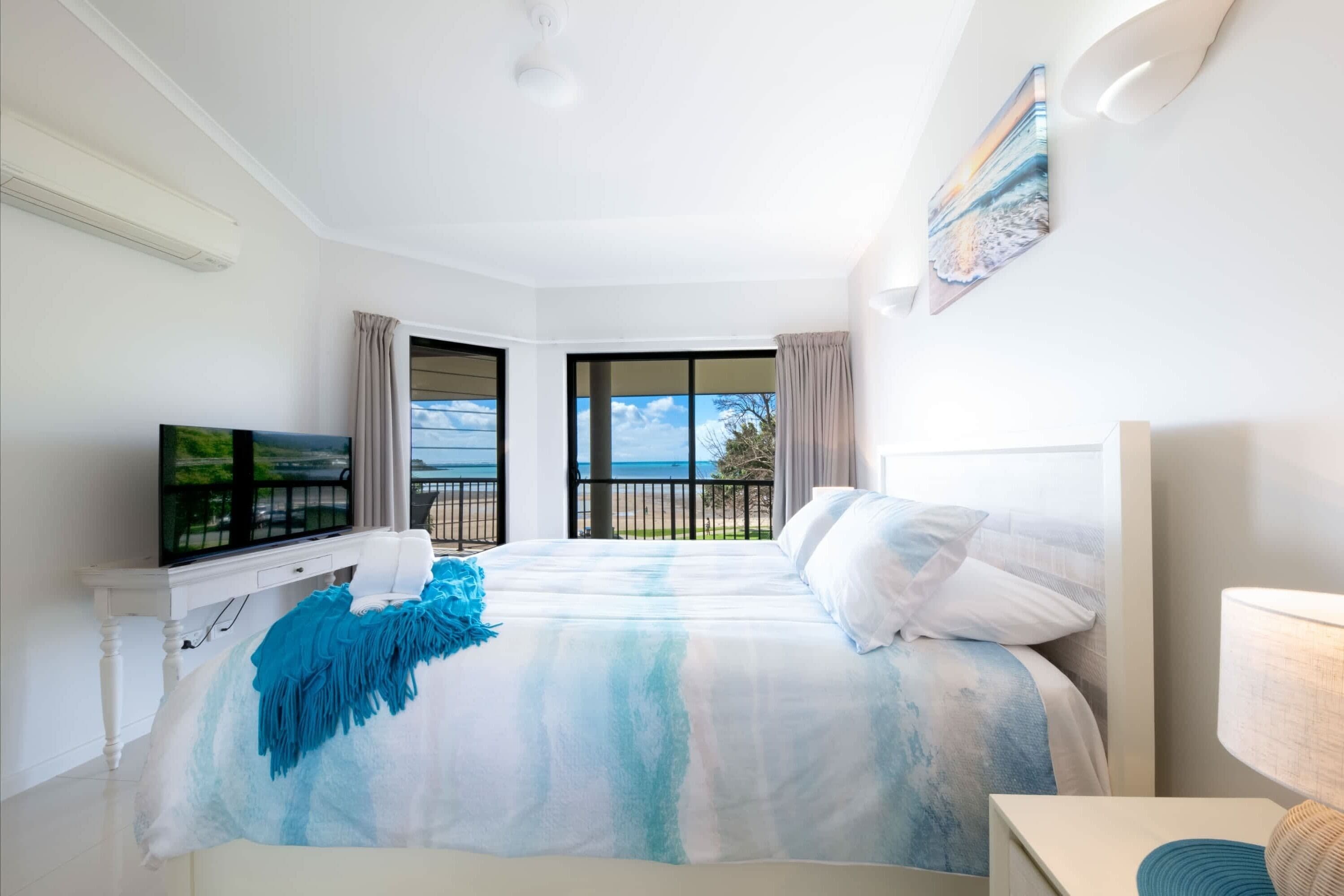 Ein Hotelzimmer mit einem großen Bett, einem Fernseher, einem Balkon mit Meerblick und einem Gemälde an der Wand.