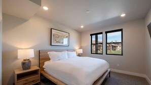 A bedroom with a bed, two bedside tables, a nightstand, a framed picture on the wall, and a window with a view. - Retreat at Jordanelle | Relaxing w/ Lake Views (Heber City)