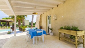 En udendørs terrasse med et bord, stole, en træbænk, en potteplante samt udsigt til en swimmingpool og grøn vegetation.