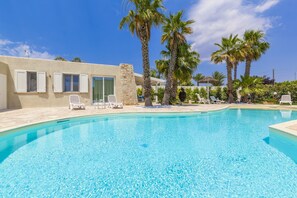 A clear blue pool with white lounge chairs, palm trees, and a building with large windows and a stone facade. - Charming villa with pool and garden (Nardò)