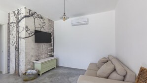En stue med et stort træ-vægmaleri, et fladskærms-tv monteret over et grønt skab, en beige sofa og en hængende lampe.