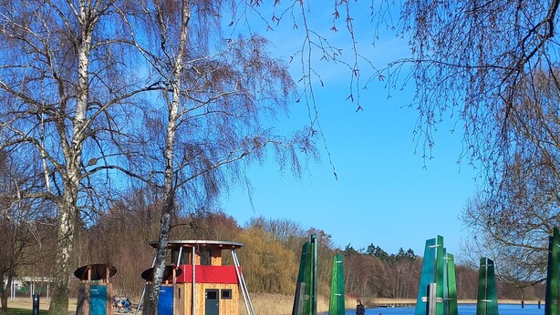 Een park met speeltoestellen, een klein houten gebouwtje en een waterpartij op de achtergrond.