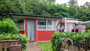Een klein, rood-wit huis met een veranda, omgeven door groen.