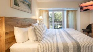 A hotel room with a wooden headboard, a large bed with striped bedding, a desk with a chair, and a balcony with curtains. - 2128 Beachwood (FERNANDINA BEACH)