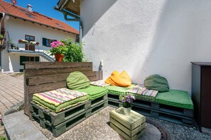 A cozy outdoor seating area with wooden benches and cushions, a potted plant, and a white wall in the background.