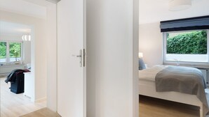 Ein Schlafzimmer mit Bett, Fenster mit Blick ins Grüne, Nachttisch mit Lampe und Tür zu einem anderen Raum.